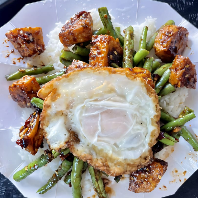 Thai dish with white rice, green beans, glazed tofu, and a fried egg on a white plate.