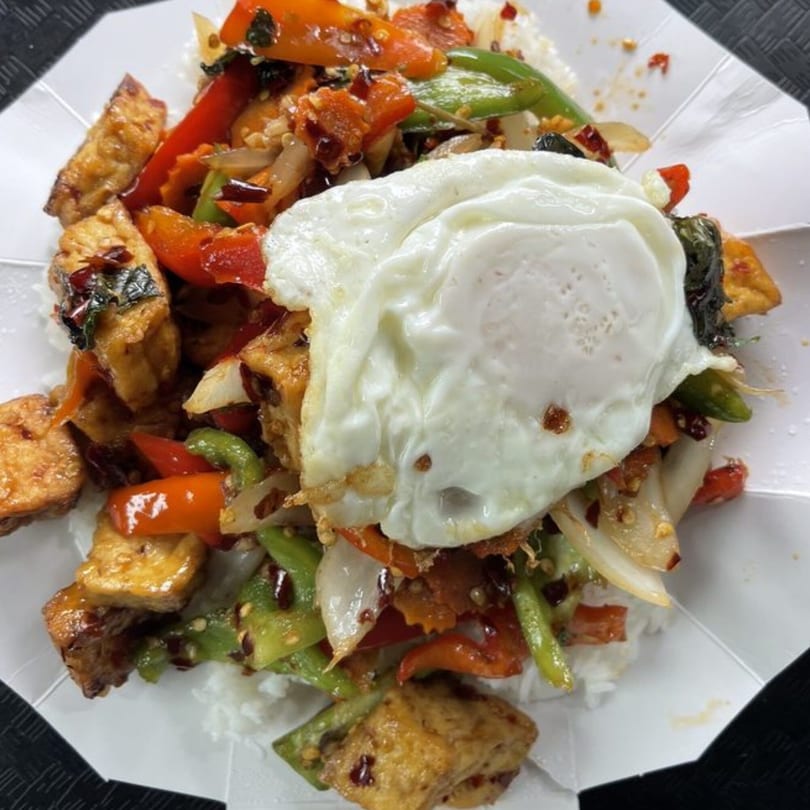 Thai basil tofu with bell peppers, onions, and a fried egg over rice in a white container.